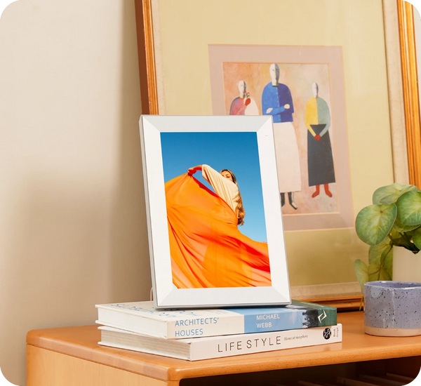 Framed photograph of a person in an orange dress on a stack of books with a plant and artwork in the background.