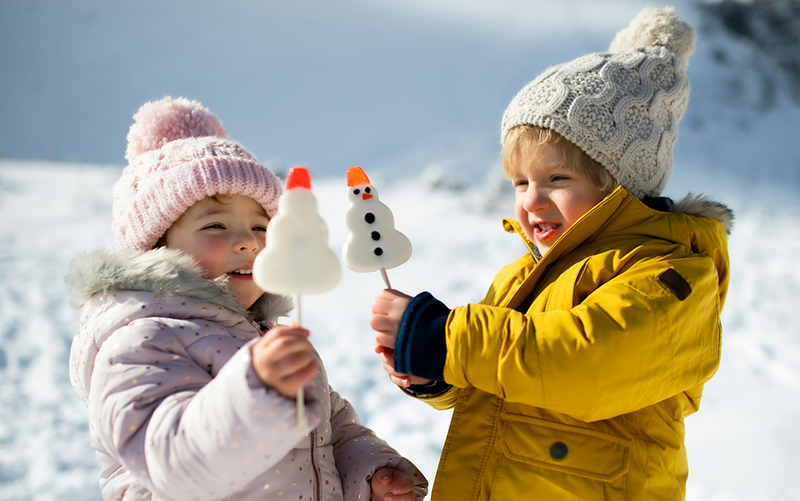 Two children in winter clothing holding snowman-shaped lollipops in a snowy landscape.