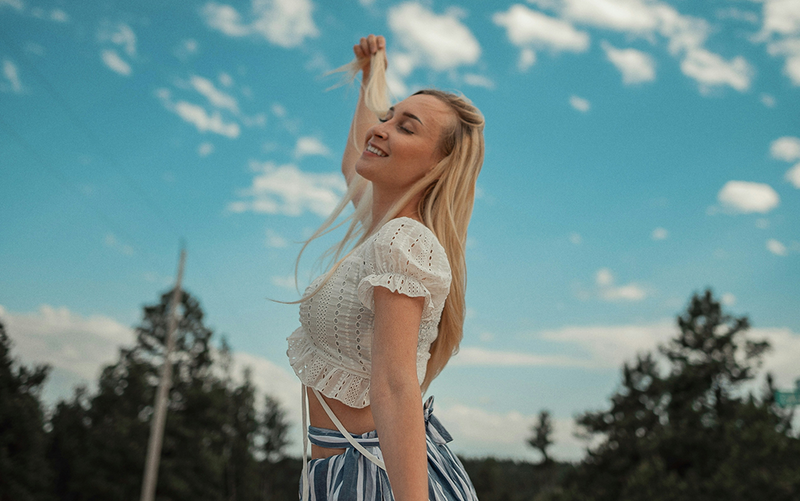 Woman in a white top and striped skirt against a blue sky with clouds and trees.