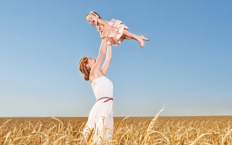 Woman in a white dress holding a child above a wheat field with a clear blue sky.