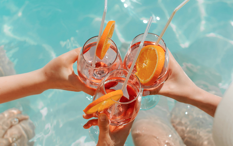 Four hands holding glasses of orange juice with straws against a blue water background