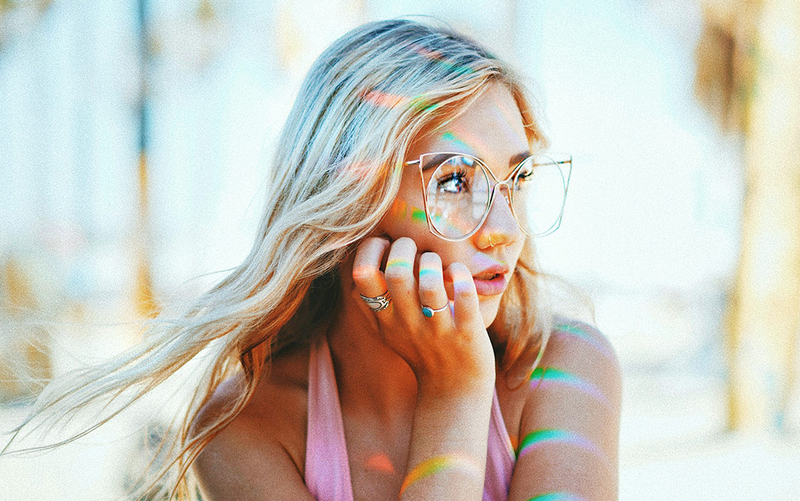Woman with colorful streaks in her hair and glasses, wearing a pink top, against a blurred natural background.